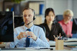 © DragonImages - Medium shot of Caucasian middle aged man wearing headset, working at call center desk with team in office. Illustrating customer support, sales service, professional business staffing