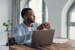 © Daniel - Young black man at a wooden desk, thoughtfully gazing away from his laptop while working remotely in a modern apartment home office, planning and concentrating on a project