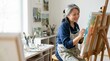 © OlgaNeuroArt - Retired Asian woman painting on canvas in her art studio. Smiling senior female artist using a paintbrush at a wooden easel. Creative hobby and retirement lifestyle concept