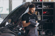 © anatoliycherkas - Mechanic examining car engine at auto repair shop, using laptop for vehicle diagnostic and maintenance