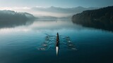 Rowers move swiftly across a calm lake in the early morning sun near mountains in the background as mist rises off the water