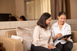 © Nalinee - A friendly young female financial advisor or caregiver sits on a living room sofa with a smiling senior Asian woman, using a digital tablet to explain information and plan together.