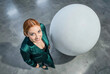 © f_bossa - Woman in emerald satin suit looks upward toward the camera from a high angle perspective beside a large white spherical object on a polished concrete floor under bright studio light.