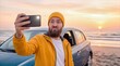 © ChaoticDesignStudio - Tourist making funny face while taking selfie with smartphone on beach at sunset. Young man posing for social media photo next to car during road trip.