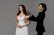 © Kaplitskaya Love - Elegant studio scene with a woman in a white strapless dress seated while a stylist in black adjusts her hair against a plain gray background with soft even