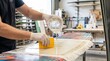 © Stitch - Surfboard shaper pouring clear liquid epoxy resin from a cup onto fiberglass cloth covering a custom board