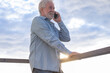 © luciano - Happy senior man with a beard enjoying a mobile phone call, standing by a wooden railing against a bright cloudy sky with sun flare, representing connection and modern technology