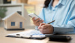 © Father_Studio - Businesswoman reviews financial documents with smartphone and pen. Her focused expression suggests careful planning. Miniature house model sits beside spreadsheets. Ideal for real estate, budgeting.