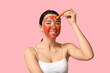 © Pixel-Shot - Beautiful young woman with facial mask using massage roller on pink background