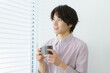 © gohan - 窓際に立つ縦縞のシャツを着てマグカップを持つ若い日本人男性のポートレート