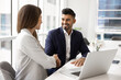 © fizkes - Smiling Indian businessman handshaking with female colleague or client seated at desk with laptop in modern office. Trust in business, cooperation and mutual understanding during business conversation