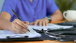 © Siwaporn - Healthcare professional reviewing medical documents and writing notes at a desk with stethoscope and laptop. Clinical documentation, patient records, healthcare administration, medical reporting