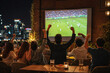 © Denkun - Group of friends cheering and drinking beer while watching a soccer match on a large outdoor screen at a bar terrace at night with city skyline lights. Urban nightlife and sports fan concept.