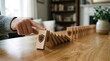 © MSTMAYA - A human hand pushing a wooden domino block with a shield causing a chain reaction on a wooden table in a blurred home office environment