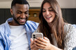 © simona - Young mixed-race couple sitting on a sofa smiling and looking at a smartphone together, sharing a moment of happiness, connection and casual home life