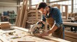 © Dara - A young man wearing safety glasses and an apron is operating a circular saw to cut a piece of wood. He is working in a workshop filled with lumber and wood panels.