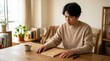 © pinchos - 温かい照明の部屋で、ノートを見つめながら穏やかに悩み、思案する表情の若い日本人男性。