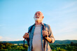 © we.bond.creations - Happy senior man smiling while holding backpack straps, enjoying active retirement outdoors under clear blue sky