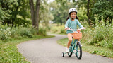 Young girl riding a bicycle with training wheels in a park. Happy child wearing a safety helmet cycling on a paved path with a doll in the basket. Outdoor summer recreation and active lifestyle