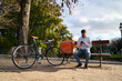 © Oscar - Delivery courier sitting on bench checking smartphone in park