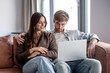 © luciano - Young couple sitting on couch and browsing internet on laptop, enjoying leisure time and togetherness
