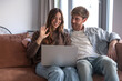 © luciano - Happy young couple networking from home, waving hand for an online video call using laptop computer