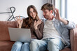 © luciano - Happy couple on sofa making video call with laptop, waving hello and smiling, connecting remotely from home