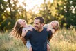 © mtmakos - A happy father giving his two laughing little daughters a piggyback ride outdoors in a beautiful sunlit field.