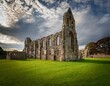 © James - ruins of a stone abbey stand in a green field under a cloudy sky evocative of history