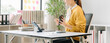 © Kritdanai - Woman Working at Desk: A woman in a yellow sweater sits at a desk in a bright office, holding a coffee cup and looking at a tablet, surrounded by books and plants.