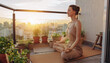 © SolaruS - Peaceful young woman practices yoga and meditation on a cozy city balcony surrounded by green plants during a warm golden sunset, embodying urban wellness and mindfulness.