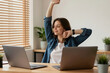 © Viktor - Young woman stretches arms up, takes break at home office desk. She feels relaxed, refreshed, energized for more work. Computer on table. Natural light indoors.