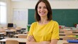 © Elena - A female teacher smiles at the camera in a classroom, standing behind desks with chairs for students. Her office and teaching materials are arranged neatly around her.