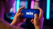 © Stocker - Man plays mobile game with word win displayed on screen, casino background with neon lights, close up of hands holding smartphone, concept of online gambling success and digital entertainment winning.