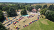© Amazing Aerial - Succieu, France - 13 June 2026: Aerial view of the Chateau de Milliassieres during a car show with rows of colorful sports cars and people on the lawn surrounded by trees and rolling hills.