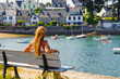 © M.studio - Back view of young female tourist sitting on wooden bench looking at traditional coastal village harbor with boats in brittany. Tour tourism in France
