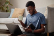 © TrueFrame Collective - Young Black man with insulin pump on arm working on laptop while holding notebook and coffee, managing home tasks. Ideal for remote work, productivity, freelance lifestyle marketing