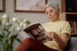 © TrueFrame Collective - Senior Asian woman reading magazine while sitting at home, showing glucose sensor on arm and independent diabetes management. Useful for healthcare campaigns, patient education