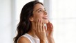 © Visionary - Beautiful young woman touching face with hands, smiling and looking up in bright room, natural skincare concept, healthy glowing skin texture