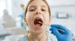© Taras Roshchuk - A young girl sits in a dental chair with her mouth open. A healthcare worker wearing blue gloves holds a swab near her throat. The clinic has modern dental equipment visible in the background