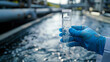 © Marina_Nov - Blue-gloved hand holds pipette filled clear water against blurred wastewater treatment plants. Scientist is testing water quality to develop environmental protection concept.