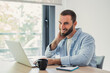 © Daniel - Smiling young man with a beard working remotely on his laptop from a bright home office, enjoying his flexible work-life balance and digital workflow