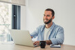 © Daniel - Young man with a beard smiling at the camera while typing on a laptop computer, enjoying remote work and a casual atmosphere at his modern home office desk with a coffee mug