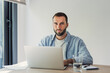 © Daniel - Young bearded man working on a laptop, sitting at a wooden desk with a notebook and glass of water, illustrating concepts of remote work and digital nomadism