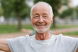 © lashkhidzetim - Old man smiles warmly outdoors. He wears light grey t-shirt. Background shows green trees and grass. Sunlight gently illuminates his face. His joyful expression suggests happiness