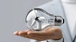 © Achirawee - A conceptual image of a light bulb with a wind turbine inside, held by a businessperson, symbolizing the idea of investing in renewable energy. The background is minimalistic, allowing for ample copy