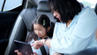 © harit - Asian mother teaching her cute little daughter with a digital tablet in the back seat of a car during a family road trip, parenting and safety travel concept.