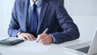 © rogerphoto - Businessman wearing suit and tie is using calculator and taking notes while working at desk in office, with laptop and stack of folders nearby, close up. Audit and taxes theme in business