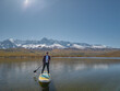 © ANDREY - Front view of man paddleboarding on scenic alpine lake against snowy mountain peaks. Active traveler on sup board exploring remote wilderness landscape under bright sunny sky.
