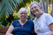 © luciano - Two happy senior adult women enjoying friendship and conversation outdoors in a tropical garden, smiling at the camera and embracing active retirement life